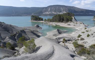 Embalse de Yesa con aguas turquesas, formaciones rocosas grises y vegetación dispersa, montañas al fondo.