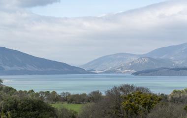 Panorámica del embalse de Yesa con montañas pirenaicas al fondo, vegetación en primer plano y cielo nublado.
