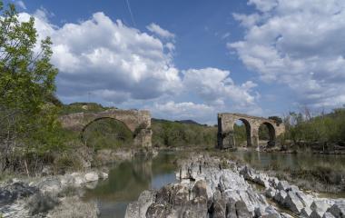 Ruins of a medieval bridge with stone arches over a river in Puente de los Roncaleses, surrounded by vegetation and mountains.