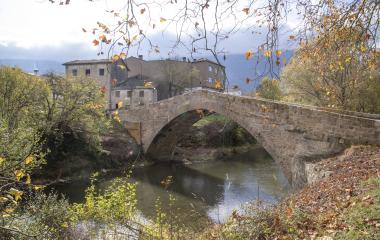 Puente de piedra medieval sobre río en Uharte Arakil con casas de piedra al fondo, rodeado de vegetación otoñal.