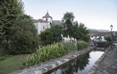 Ur-kanala eta landaredi tropikala lehen planoan, atzeko zuhaitzen artean eliza duela Urdazubin.