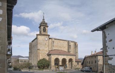 Iglesia de piedra con campanario y reloj en plaza de Urdiain, rodeada de casas tradicionales de piedra.