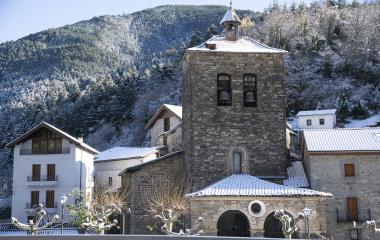 Iglesia de piedra con campanario en Urzainqui rodeada de bosque nevado y casas tradicionales de montaña.
