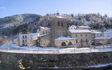 Iglesia de piedra con campanario en Urzainqui, rodeada de nieve con bosque pirenaico y río en primer plano.