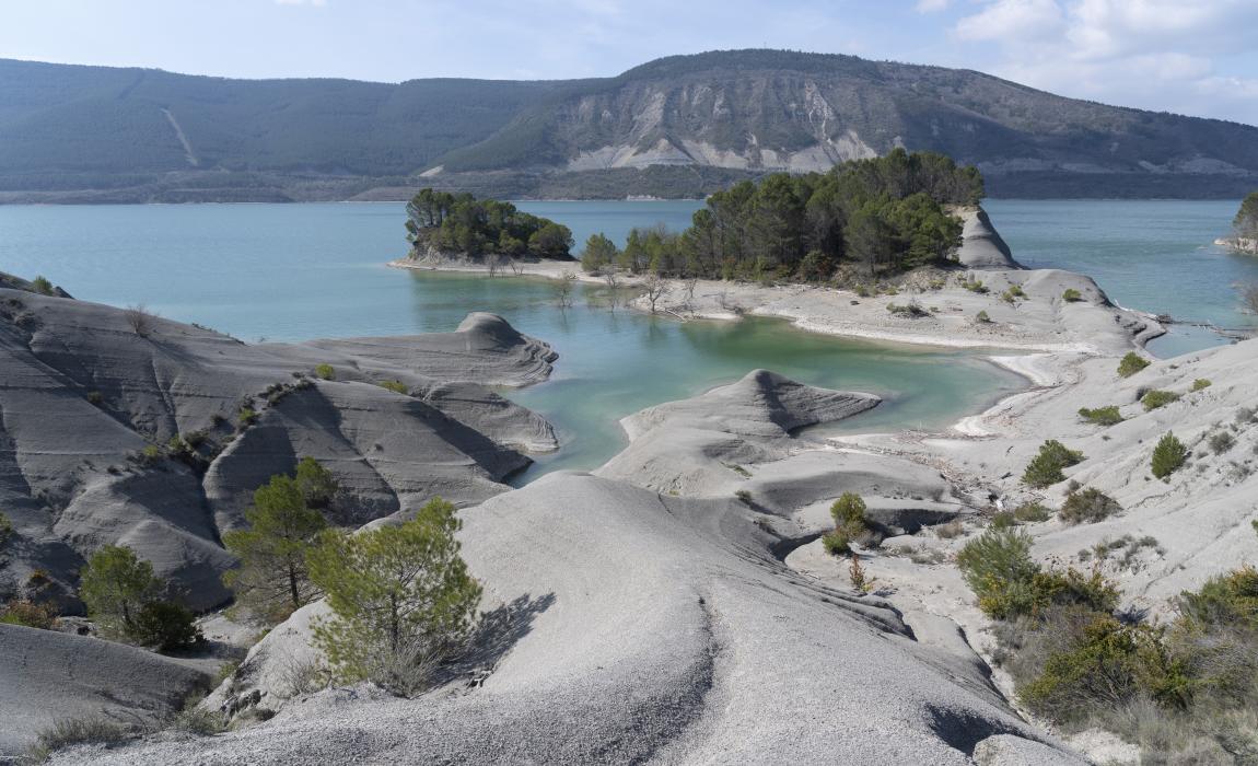 Pantano de Yesa con azul turquesa, piedras, una islita y montes al fondo.